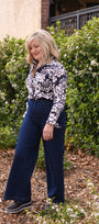 Woman in a black and white patterned top and blue pants standing outdoors with greenery in the background