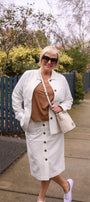 Woman in a white coat and skirt with a brown top, standing on a sidewalk.