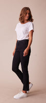 A woman modeling black slim-fit jeans with a white t-shirt and white sneakers, standing against a solid beige studio background.