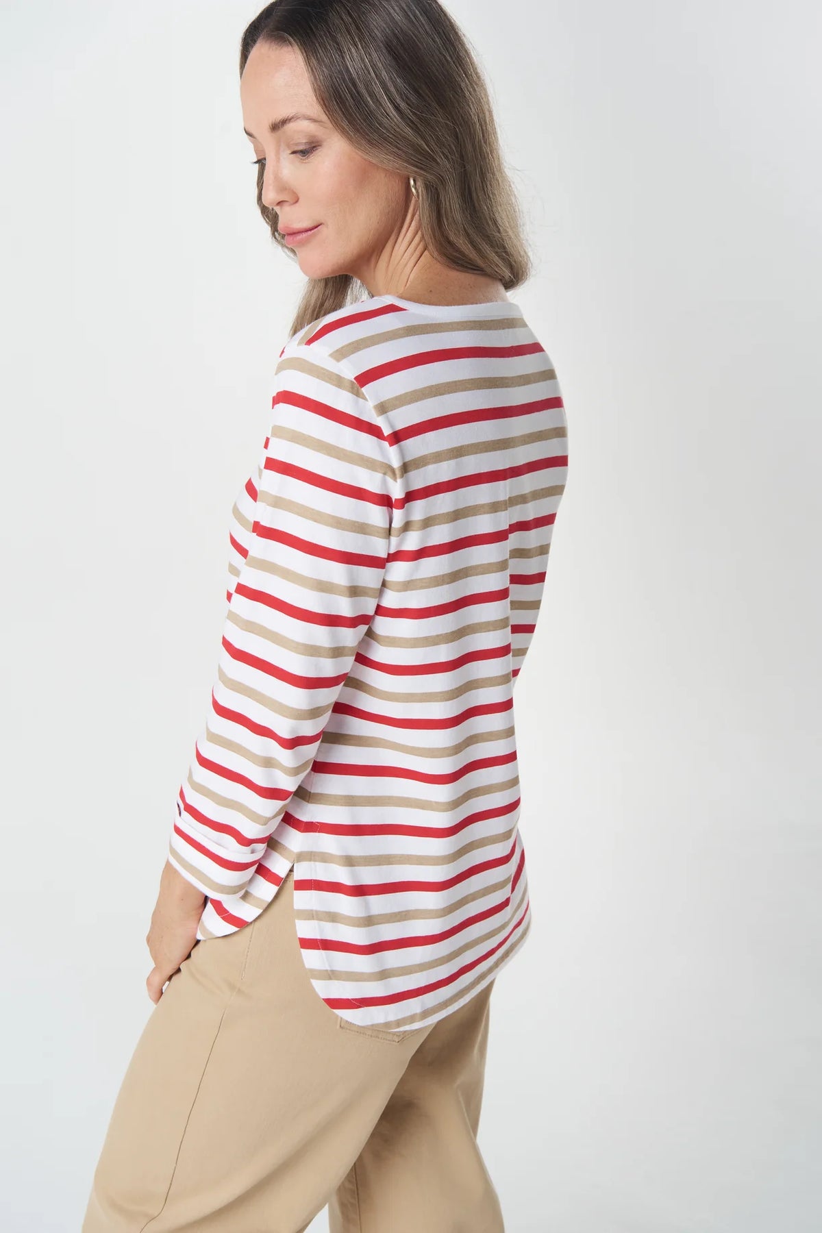 A woman wearing a long-sleeve striped top with red, beige, and white horizontal stripes, paired with beige pants, shown from a side-back angle against a light gray studio background.