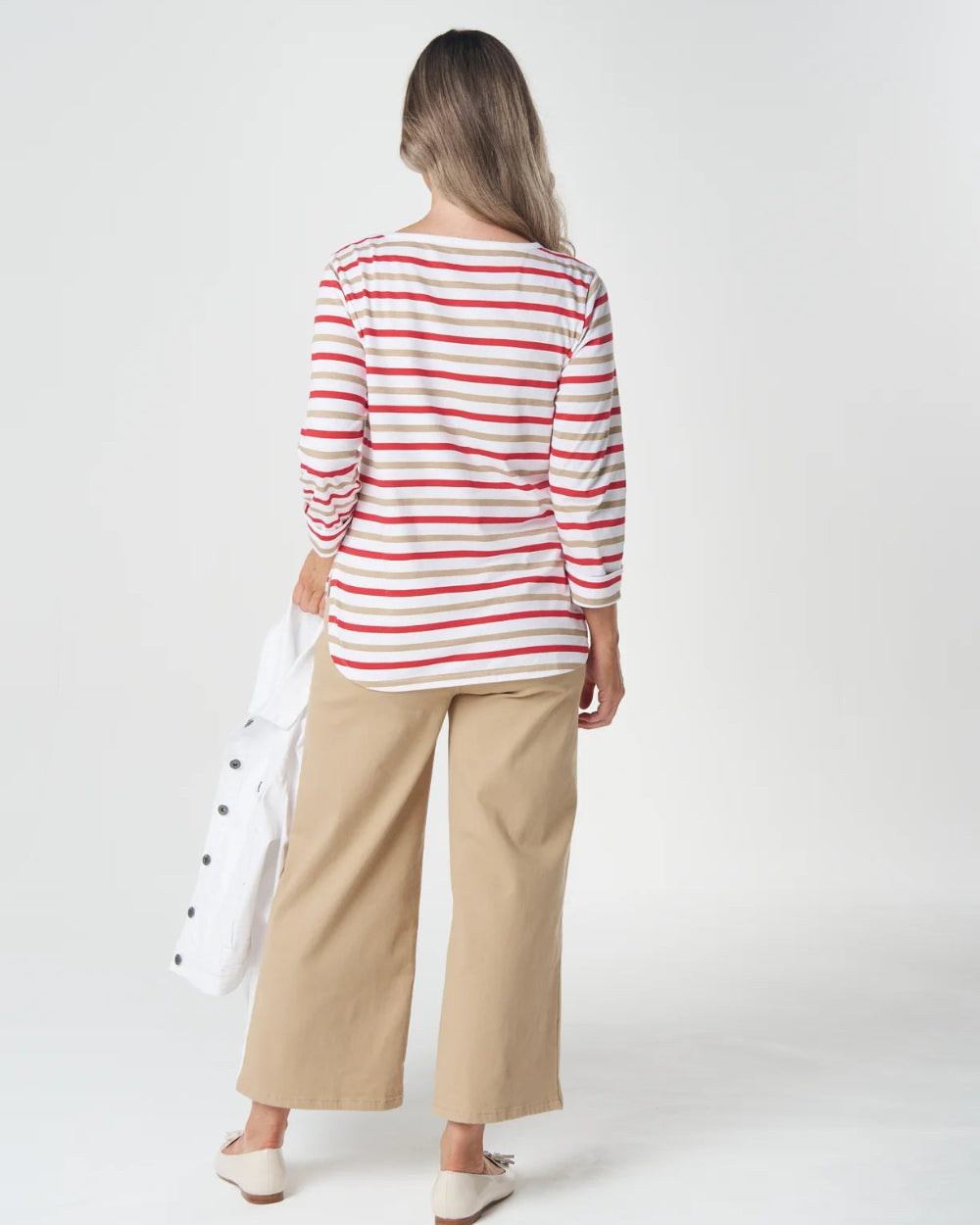 A woman is shown from behind wearing a red and white striped long-sleeve top, beige wide-leg cropped pants, and white shoes, holding a white bag against a plain white studio background.