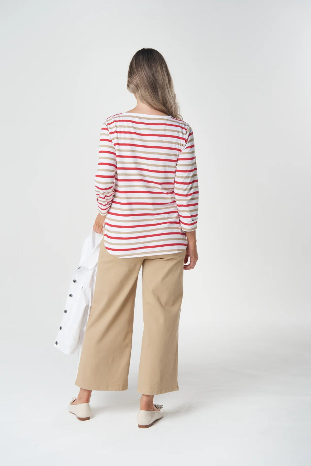 A woman is shown from behind wearing a red and white striped long-sleeve top, beige wide-leg cropped pants, and white shoes, holding a white bag against a plain white studio background.