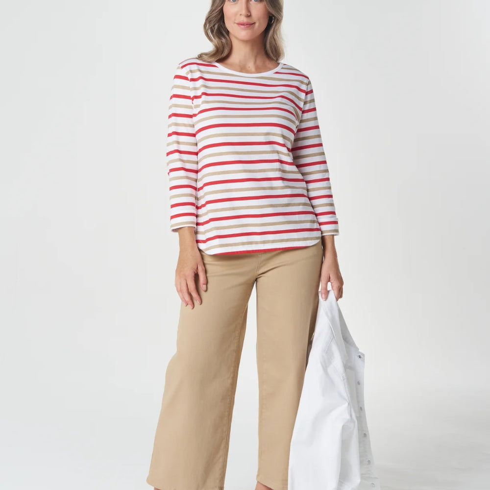 A woman modeling a red and white horizontally striped long-sleeve top with beige cropped trousers, holding a white jacket against a light gray studio background.