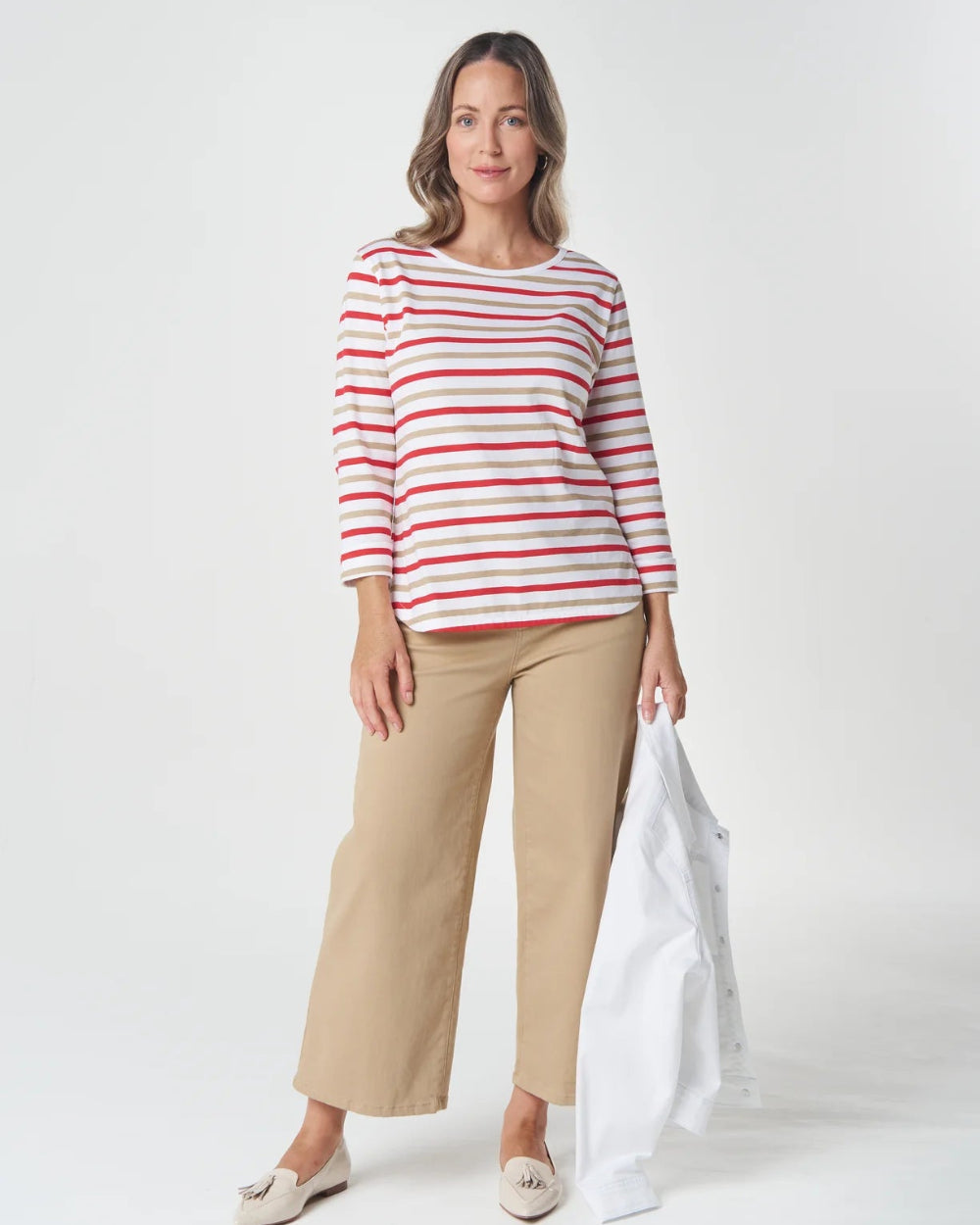 A woman modeling a red and white horizontally striped long-sleeve top with beige cropped trousers, holding a white jacket against a light gray studio background.