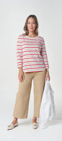 A woman modeling a red and white horizontally striped long-sleeve top with beige cropped trousers, holding a white jacket against a light gray studio background.