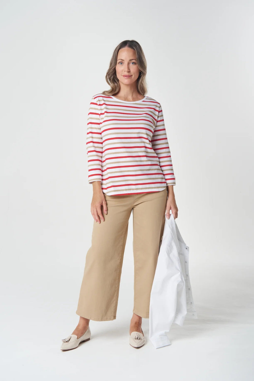 A woman modeling a red and white horizontally striped long-sleeve top with beige cropped trousers, holding a white jacket against a light gray studio background.