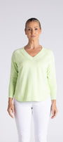 A woman modeling a light lime green long-sleeve V-neck knit top with white pants, standing against a light gray studio background.