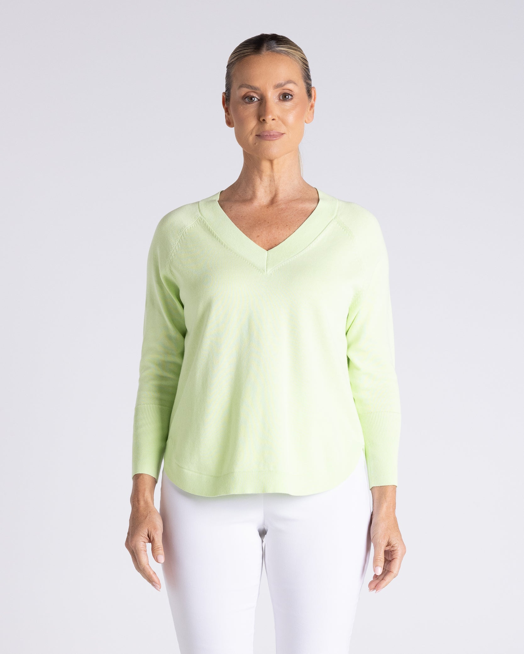 A woman modeling a light lime green long-sleeve V-neck knit top with white pants, standing against a light gray studio background.