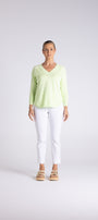 A woman stands against a light gray studio background wearing a pale yellow-green long-sleeve V-neck top, white cropped pants, and tan sandals.