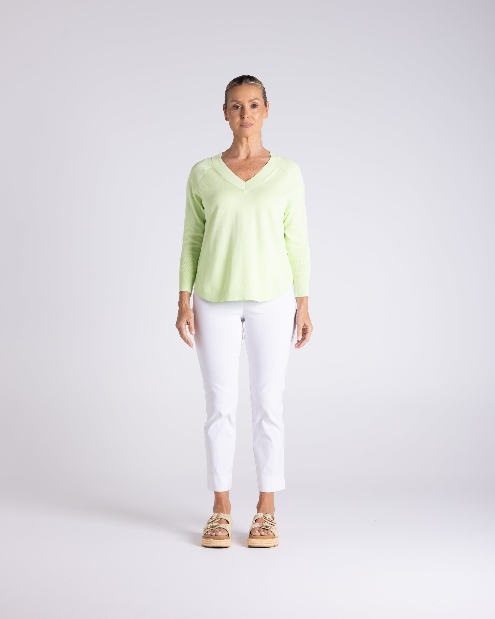 A woman stands against a light gray studio background wearing a pale yellow-green long-sleeve V-neck top, white cropped pants, and tan sandals.