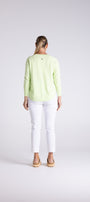 A woman viewed from behind wearing a light lime green long-sleeve top and white cropped jeans, standing against a light gray studio background.