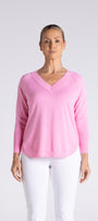 A woman modeling a light pink long-sleeve V-neck sweater with white pants, standing against a light gray studio background.
