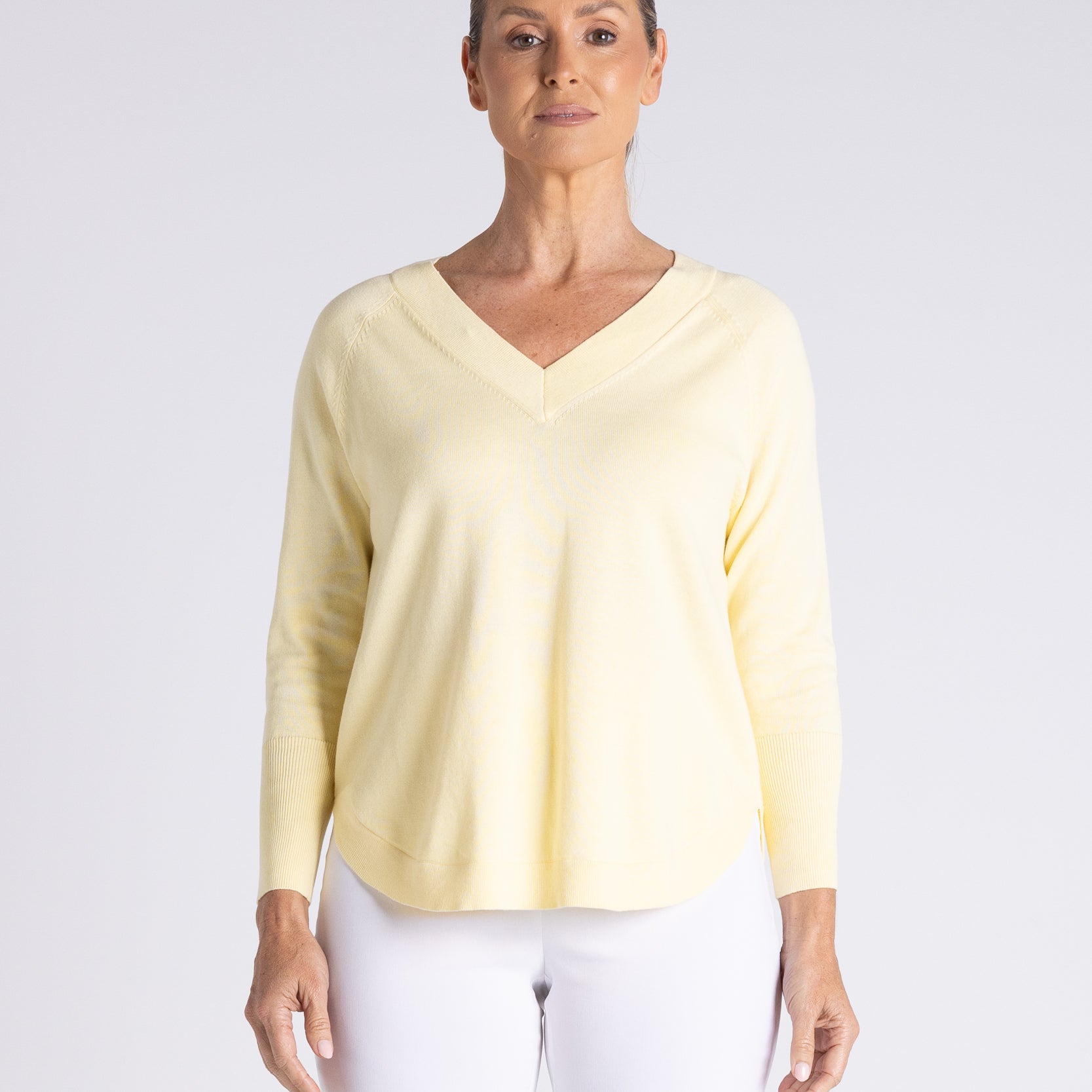 A woman modeling a pale yellow long-sleeve V-neck top with white pants, standing against a light gray studio background.