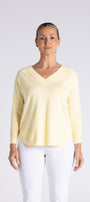 A woman modeling a pale yellow long-sleeve V-neck top with white pants, standing against a light gray studio background.