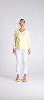 A woman stands against a light gray studio background wearing a pale yellow long-sleeve V-neck top, white cropped pants, and tan sandals.