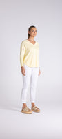 A woman modeling a pale yellow long-sleeve V-neck top with white cropped pants and tan sandals, standing against a light gray studio background.