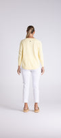 A woman viewed from behind wearing a pale yellow long-sleeve top, white cropped pants, and tan sandals, standing against a light gray studio background.