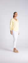 A woman stands in profile wearing a pale yellow long-sleeve V-neck top, white pants, and woven sandals against a light gray studio background.
