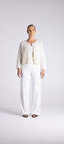 A woman stands against a light gray studio background wearing a white three-piece outfit consisting of a textured short cardigan, a fitted top, and wide-leg trousers.