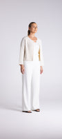 A woman stands in a studio wearing a white textured cardigan, white wide-leg pants, and a light gold top. She is photographed against a seamless light gray background.