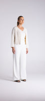 A woman modeling a white wide-leg pantsuit with a textured cream cardigan and a gold inner top, standing against a light gray studio background.