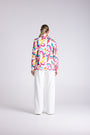 A woman viewed from behind wearing a long-sleeve button-up shirt with a vibrant multicolor abstract pattern and white wide-leg trousers, set against a plain light gray studio background.