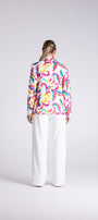 A woman viewed from behind wearing a long-sleeve button-up shirt with a vibrant multicolor abstract pattern and white wide-leg trousers, set against a plain light gray studio background.