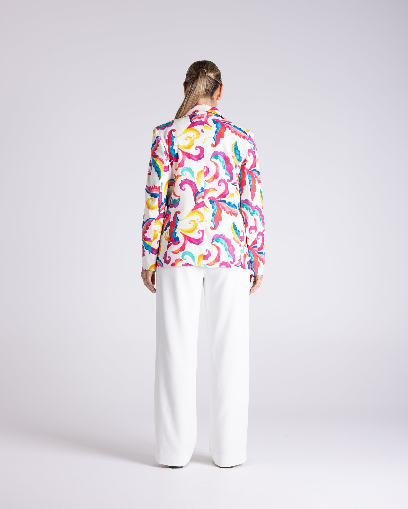 A woman viewed from behind wearing a long-sleeve button-up shirt with a vibrant multicolor abstract pattern and white wide-leg trousers, set against a plain light gray studio background.