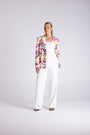 A woman modeling a white wide-leg jumpsuit with a colorful abstract print blazer, standing against a light gray studio background.