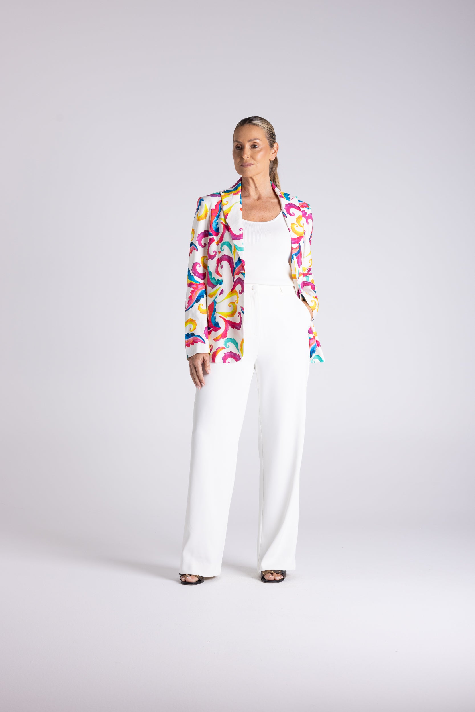 A woman modeling a white wide-leg jumpsuit with a colorful abstract print blazer, standing against a light gray studio background.