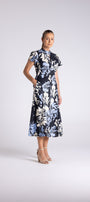 A woman modeling a short-sleeved midi dress with a navy blue base and a large white and light blue floral print, standing against a light gray studio background.