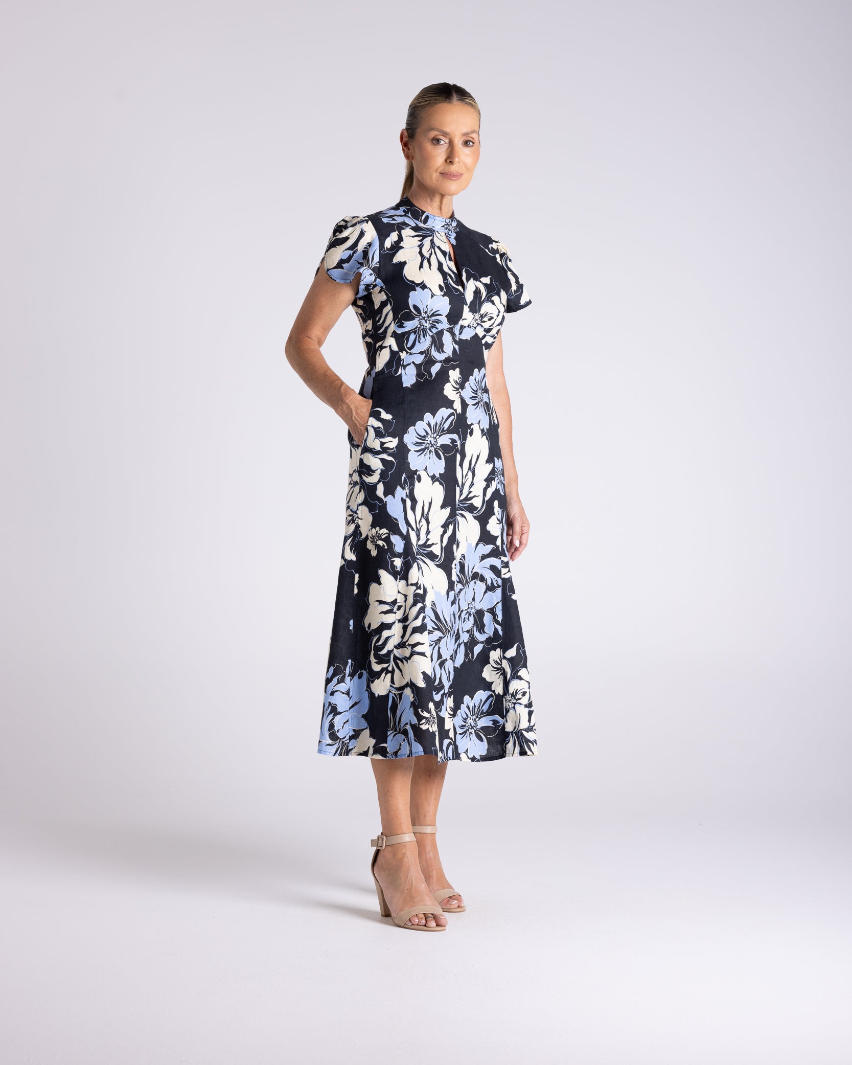 A woman modeling a short-sleeved midi dress with a navy blue base and a large white and light blue floral print, standing against a light gray studio background.