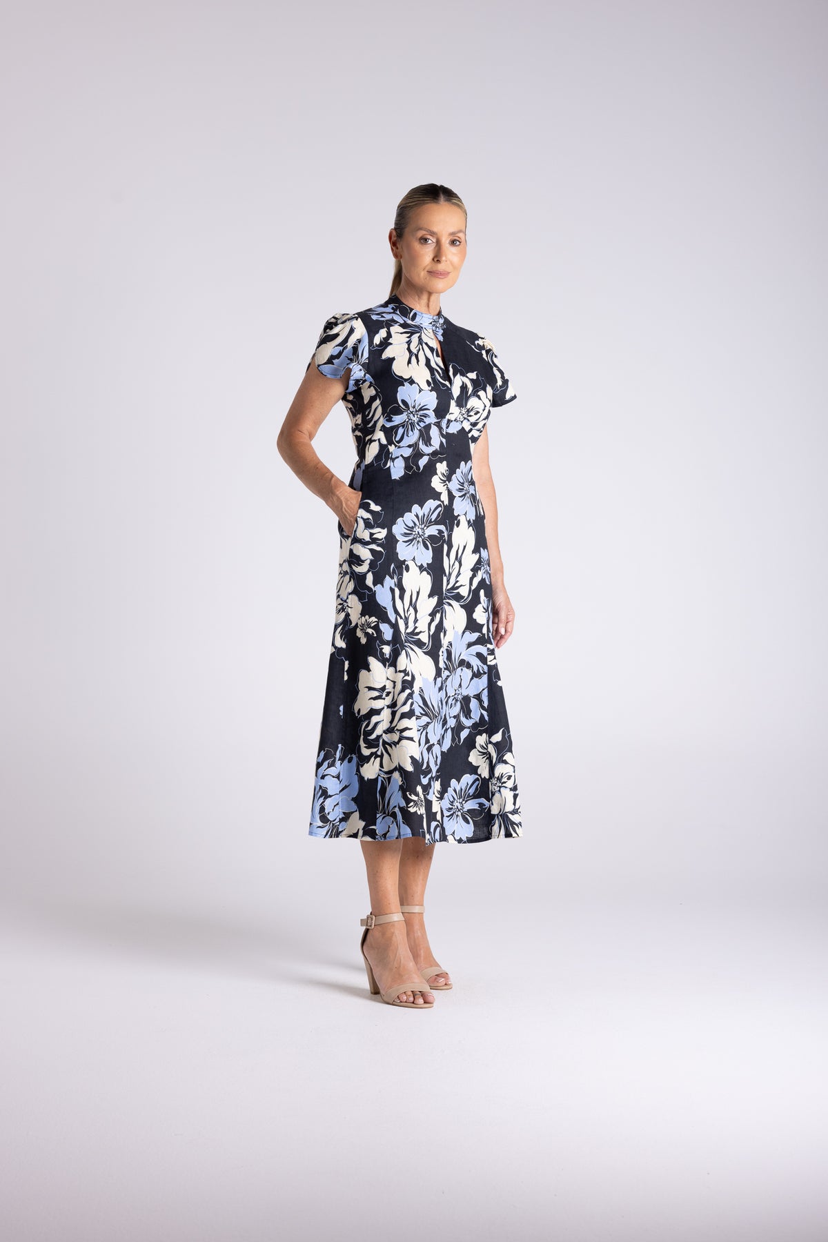 A woman modeling a short-sleeved midi dress with a navy blue base and a large white and light blue floral print, standing against a light gray studio background.