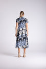 A woman viewed from behind wearing a short-sleeve midi dress with a black base and a large light blue and white floral print, paired with beige high-heeled sandals, against a light gray studio background.