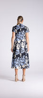 A woman viewed from behind wearing a short-sleeve midi dress with a black base and a large light blue and white floral print, paired with beige high-heeled sandals, against a light gray studio background.