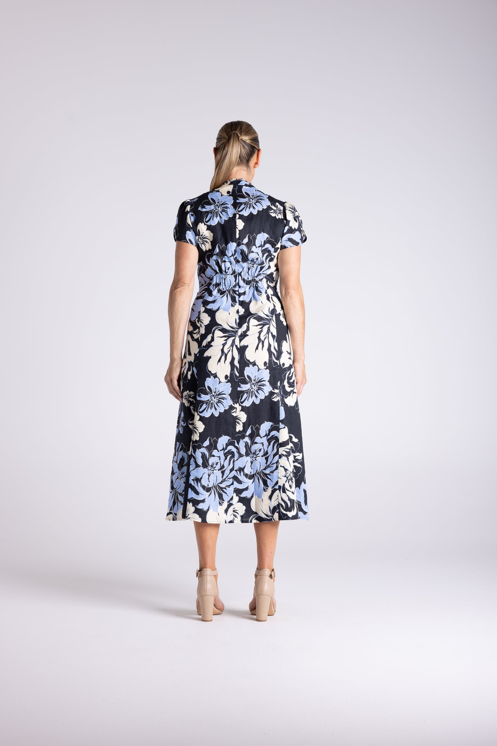 A woman viewed from behind wearing a short-sleeve midi dress with a black base and a large light blue and white floral print, paired with beige high-heeled sandals, against a light gray studio background.