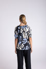 A woman viewed from the back wearing a short-sleeve blouse with a navy, cream, and light blue floral pattern, paired with dark pants, against a light gray studio background.