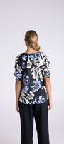 A woman viewed from the back wearing a short-sleeve blouse with a navy, cream, and light blue floral pattern, paired with dark pants, against a light gray studio background.