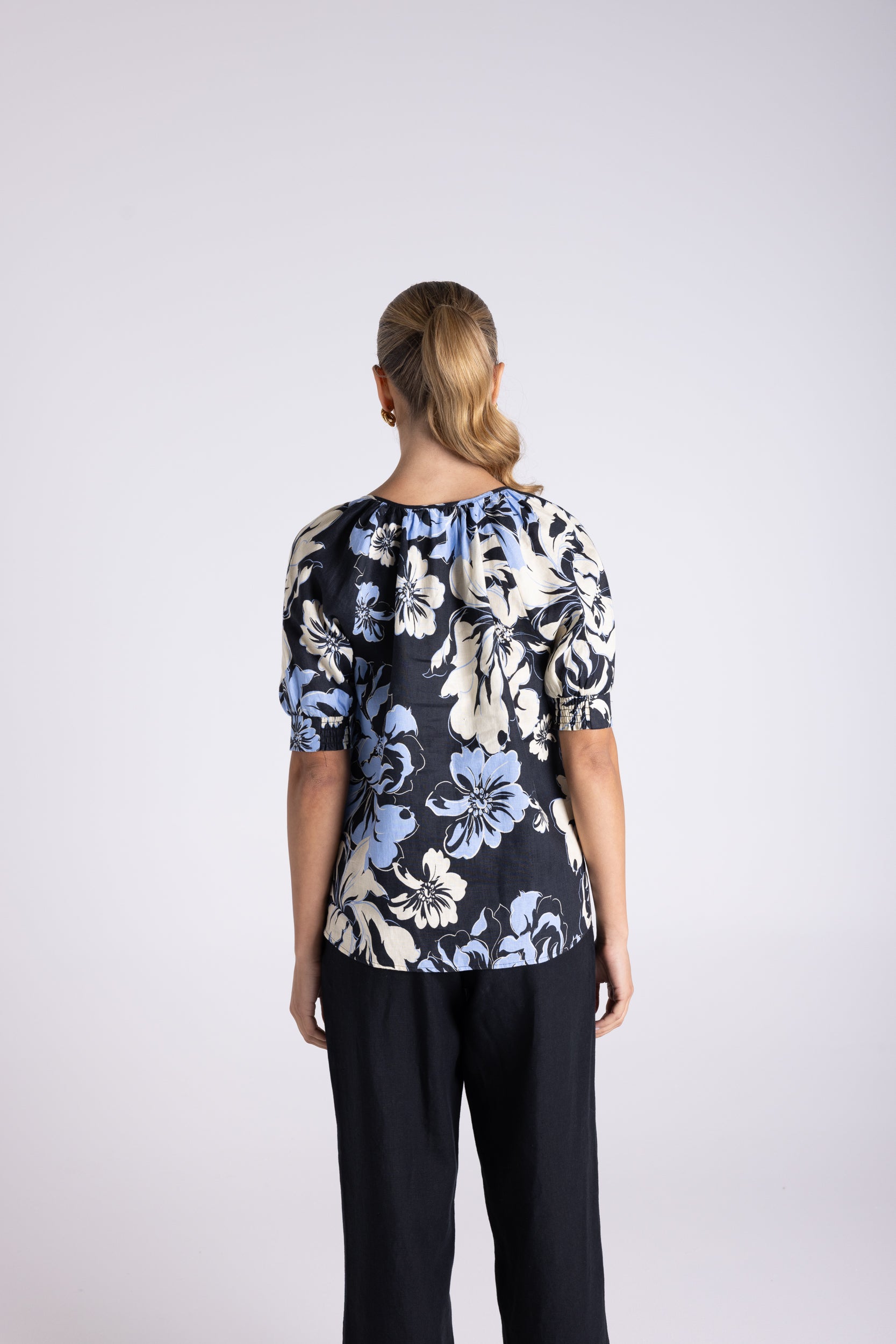 A woman viewed from the back wearing a short-sleeve blouse with a navy, cream, and light blue floral pattern, paired with dark pants, against a light gray studio background.