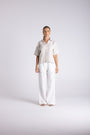 A woman stands against a light gray studio background wearing a short-sleeve light beige button-up shirt and white wide-leg pants.