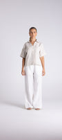 A woman stands against a light gray studio background wearing a short sleeve light beige button up shirt and white wide leg pants.