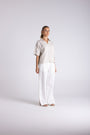 A woman modeling a light beige short-sleeve button-up shirt paired with white wide-leg trousers, standing against a light gray studio background.