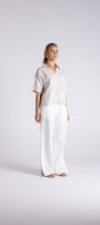 A woman modeling a light beige short-sleeve button up shirt paired with white wide leg trousers, standing against a light gray studio background.
