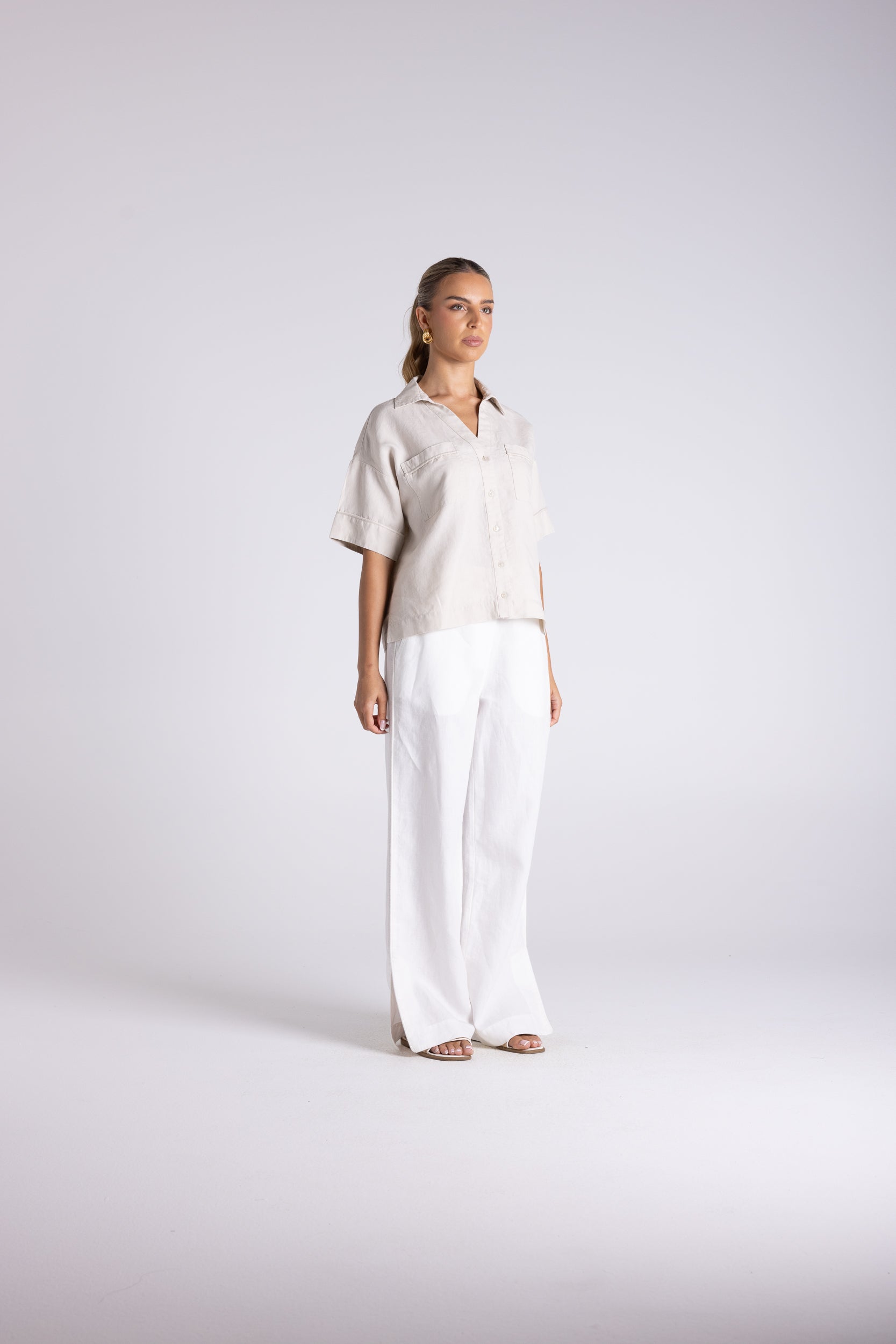 A woman modeling a light beige short-sleeve button-up shirt paired with white wide-leg trousers, standing against a light gray studio background.