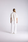 A model viewed from behind wearing a short-sleeve cream linen shirt and matching wide-leg linen trousers against a light gray studio background.