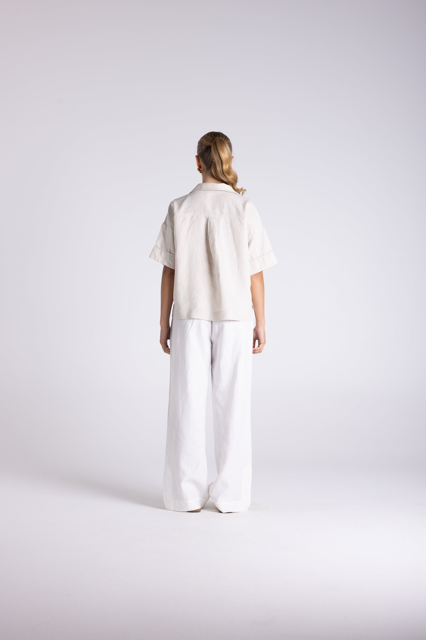 A model viewed from behind wearing a short-sleeve cream linen shirt and matching wide-leg linen trousers against a light gray studio background.