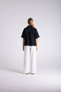 A model viewed from the back wearing a short-sleeved black top and wide-leg white trousers, standing against a light gray studio background.