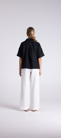 A model viewed from the back wearing a short sleeved Navy top and wide leg white trousers, standing against a light gray studio background.