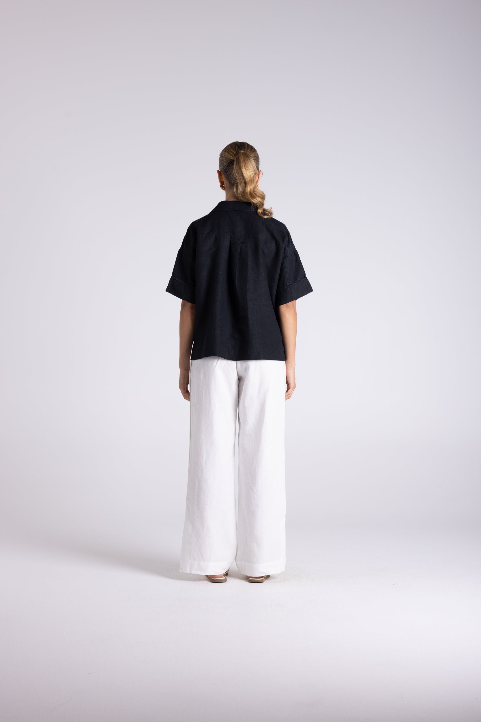 A model viewed from the back wearing a short-sleeved black top and wide-leg white trousers, standing against a light gray studio background.