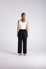 A woman standing with her back to the camera, wearing a white sleeveless top and black wide-leg trousers against a light gray studio background.